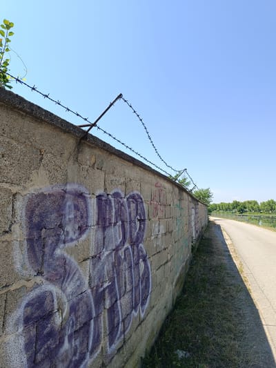 The image depicts a concrete block wall with graffiti on its surface, topped with barbed wire. To the right of the wall is an asphalt path running alongside a body of water, likely the Jiu River, bordered by trees. The sky is clear and blue. This scene is located in Târgu Jiu, Romania, near the Jiu River, a common area for walking and recreation.