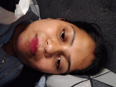 The image shows a woman lying on a bed, with her face and upper body visible in the frame. She is wearing a blue shirt with a silver chain necklace. Her eyes are closed, and she is wearing red lipstick. Her dark hair is spread across the pillow. The room has a black and gray, patterned carpet. The image was taken in Schwarzenbek, Germany, at coordinates 53.509, 10.492.