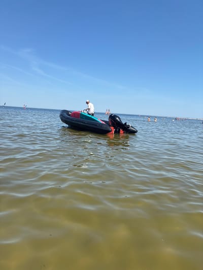 The image shows a black inflatable boat with a red and teal interior, equipped with an outboard motor, floating in calm, shallow, greenish water. A person wearing a white cap and shirt stands in the boat, holding the steering wheel. In the background, several people are visible in the water, some swimming and others engaged in water sports such as windsurfing. The sky is clear blue with a few wispy clouds. The scene is consistent with a beach or waterfront area in Rewa, Poland, a location known for its shallow waters and water sports activities.
