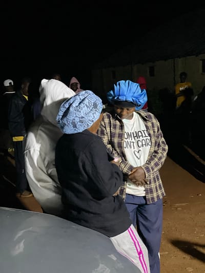 In Wumat, Nigeria, at night, a group of individuals are gathered outdoors near a building. Two women are in the foreground, both wearing bonnets. The woman on the right, wearing a blue bonnet, is dressed in a plaid shirt over a white t-shirt with the text "I DON'T CARE" and a heart symbol. She is looking down. The woman on the left, wearing a light blue lace bonnet, faces away from the camera and is dressed in a black jacket. A person in a white hooded garment is positioned between them. Other individuals are visible in the background, including one in a yellow shirt. The ground appears to be dirt.