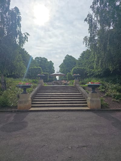 The image depicts a wide shot of a formal garden pathway leading up a grand stone staircase. The pathway is paved with dark asphalt, which transitions into a wide set of stone steps. Flanking the staircase are stone planters filled with colorful flowers. The stairs ascend to a landscaped garden area with manicured hedges and trees. In the distance, at the top of the stairs, a decorative gazebo with an ornate roof is visible. The sky above is overcast with the sun shining through, casting a lens flare effect on the left side of the image. The overall scene suggests a well-maintained public park or botanical garden. This image is of the steps and gazebo at the University Hospital of North Tees in Stockton-on-Tees, United Kingdom.