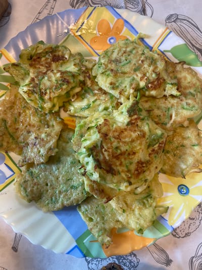 The image shows a plate of zucchini fritters on a patterned tablecloth. The fritters are golden brown and appear to be made with shredded zucchini and batter. They are arranged in a pile on a colorful, scalloped-edge plate with a floral design. The tablecloth has a greyish-white background with black line drawings of vegetables. The scene is likely indoors, possibly in a home kitchen. There are no people or animals visible in the image. The location is not specified in the image, and therefore, it cannot be confirmed to be in the city of Турово, Russia.
