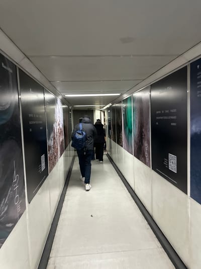 The image shows a pedestrian walkway, possibly in Pudahuel, Chile, based on the Spanish text "DENTRO DE CHILE PUEDES" (inside Chile you can). The walls are lined with posters featuring text and images, some with QR codes. A person with a dark jacket, blue backpack, and dark pants is walking away from the camera through the walkway. The walkway is well-lit and has a neutral color scheme with a mix of white and dark gray. The perspective makes it appear as if the walkway is diminishing into the distance.