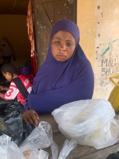 The image depicts an outdoor market or shop entrance in Okene, Nigeria. In the foreground, an adult female with a neutral expression, wearing a dark purple hijab, looks directly forward from behind a weathered wooden counter. Her right hand rests on the counter. Displayed on the counter are multiple clear plastic bags, one containing light-colored goods, and a black plastic bag. To the left, a younger female with braided hair, seen from the back, wears a red and white shirt displaying 'ALEXANDR' and is engaged in an activity within the dimly lit interior. Another person in a light hijab is partially visible further inside. A weathered wooden door, marked with numbers (e.g., '08', '0312', '53'), frames this interior. To the right, a stained, light yellow exterior wall features green graffiti spelling 'MALL SHU' and other faint markings. A yellow sack is partially visible on the far right.