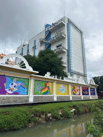 This media file shows a building with several banners celebrating Phuket City Pride. The banners feature rainbow flags, a rabbit character wearing a rainbow bowtie, and text in English and Thai. Some of the visible text includes "PHUKET CITY PRIDE 2026", "27 JUNE 2026", and "Discover Phuket". The building appears to be multi-story and is decorated with white ornamental features. The scene is set outdoors, with a green grassy area and a body of water to the right of the building. Trees are visible behind and to the left of the building. The sky is overcast, suggesting a cloudy day. The banners suggest an upcoming Pride event in 2026. The overall impression is one of festive preparation for a community event.