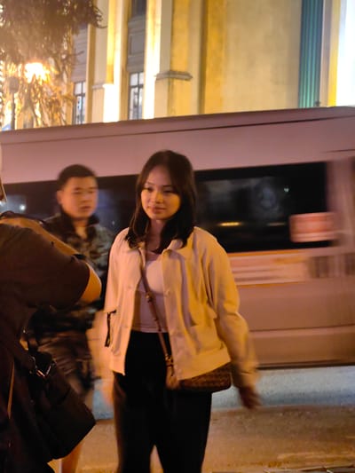 This image captures a nighttime street scene in Sa Pa, Vietnam, illuminated by artificial light sources. The central figure is a young adult East Asian female, positioned slightly right of center, looking slightly to her right with a faint smile. She is wearing a light-colored, possibly beige or off-white, jacket over a white top, and dark trousers. A small, light-brown shoulder bag with a patterned flap hangs across her body. Her right arm shows minor motion blur.
Behind and to the left of the central figure, a male of East Asian ethnicity is visible, wearing a dark, possibly camouflage-patterned jacket. He is looking forward and is slightly out of focus. On the far left, a third individual's left torso and arm are partially visible, wearing dark clothing and holding a dark bag with a lighter metal clasp.
In the mid-ground, directly behind the individuals, a light-colored vehicle, likely a bus or large van, is in motion, indicated by prominent horizontal motion blur across its body and windows. The background features a large, well-lit building with multiple rectangular windows and distinct architectural columns, presenting a classical or colonial style. On the far left, a street lamp emitting warm yellow light is visible, partially obscured by dark tree foliage. The ground beneath the figures appears to be a paved surface.