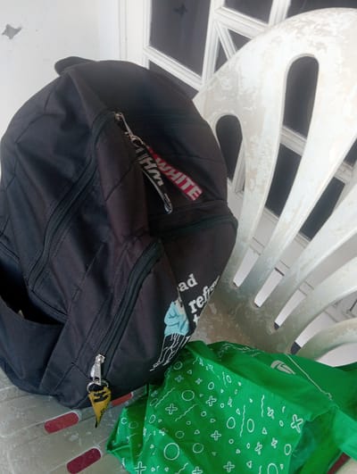 A black backpack with a red and white tag hangs from a white plastic chair. A green bag with a white geometric pattern is in front of the backpack. The objects are on a grey surface with red stripes. The background shows a white door with glass panels. The scene is likely outdoors, possibly in an urban setting.