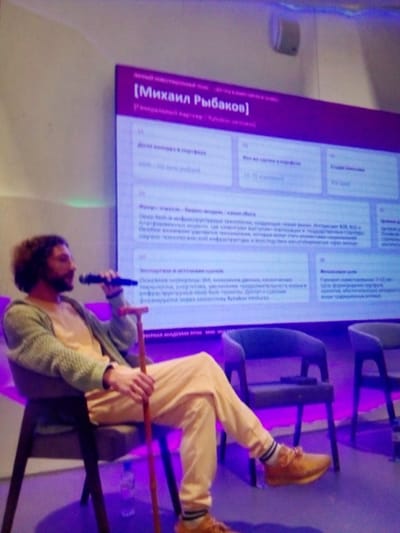 A man with curly dark hair and a beard, wearing glasses, a light-colored shirt, a knitted cardigan, and light-colored pants with striped socks and tan shoes, is seated on a chair. He is holding a microphone in his right hand and a wooden cane in his left hand. The man is looking towards the right. Behind him is a large screen displaying text in Russian, with the name "[Михаил Рыбаков]" prominently displayed. The screen is illuminated with purple light, which also casts a purple glow on the surrounding environment, including two chairs to the man's right and the floor. The setting appears to be an indoor event or conference in посёлок дачного хозяйства «Архангельское», Russia.