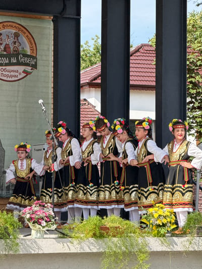A group of eight young girls dressed in traditional Bulgarian folk costumes stand on a stage. The costumes feature white blouses, black vests adorned with gold embroidery and colorful trim, and black skirts with elaborate gold patterns. Each girl wears a floral headband. They are arranged in two rows and appear to be performing a dance. In the foreground, there are two bouquets of flowers and lush green foliage. A microphone stands on a stand behind the girls, and a banner with Bulgarian text and imagery is visible on the left side of the stage. The background shows buildings with red tiled roofs and green trees, under a bright blue sky. The banner on the left indicates the event is related to a folklore competition in Pordim, Bulgaria.