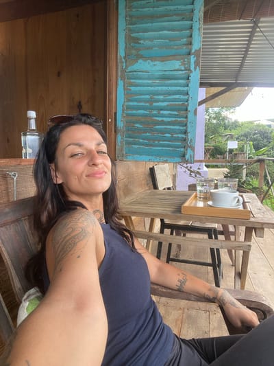 A woman is lying on a wooden deck, her head resting on her arm. She has long, dark hair and is wearing a dark blue tank top. She has tattoos on her arms. She is looking up at the camera with a slight smile.

In the background, there is a wooden table with two glasses and a white teapot. Beyond the table, there is a light purple wall and lush greenery, suggesting an outdoor setting. The overall aesthetic is rustic and relaxed. The time of day appears to be daytime, with natural light illuminating the scene.

There is a small, clear bottle with a white cap on the wooden surface in front of the woman. The scene is set in Thôn 3, Vietnam, a detail that provides geographical context. The woman's expression and the relaxed pose suggest a leisurely moment.