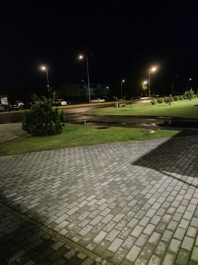 The image depicts a street scene at night in the city of Pabradė, Lithuania. The foreground features a paved walkway made of interlocking grey bricks, with some areas appearing wet, suggesting recent rain. A dark shadow stretches across the walkway from the right side of the frame. Beyond the walkway, a patch of green grass is visible, with a small, dense evergreen bush in the center.
Further into the scene, a paved road runs horizontally, with parked cars lining the left side. Streetlights illuminate the area, casting a warm glow on the road and surrounding grass. Several tall streetlights with visible bulbs are present. In the background, buildings with illuminated windows are visible, hinting at an urban or suburban environment. The overall atmosphere is quiet and dimly lit, characteristic of a nighttime setting in a Lithuanian city.