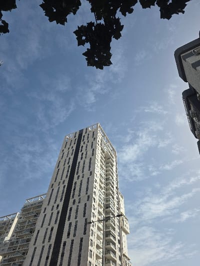 A low-angle, daytime photograph depicts a modern urban scene in Rishon LeZion, Israel, specifically within the City Center (Lev Ha'Ir) development area. The central subject is a tall, slender, light-colored multi-story building, characterized by a facade composed of numerous rectangular windows and prominent dark vertical paneling. Balconies are visible along the right side of this structure. To its immediate left, a similar, slightly recessed building with balconies is partially visible. On the far right edge of the frame, a segment of a darker, blocky building with a flat top is present. A black utility pole or streetlight, equipped with horizontal arms, extends into the lower right foreground, partially obscuring the main building. The upper portion of the image is dominated by a clear blue sky with scattered, wispy white clouds. Dark, silhouetted tree foliage frames the top of the photograph, indicating an upward perspective from beneath a tree canopy. No individuals or discernible activities are visible. The scene is illuminated by bright daylight.