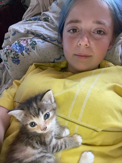 The image shows a young person with blue-dyed hair lying down, with a small, tabby kitten on their chest. The person's face is visible, and they are looking towards the camera with a gentle expression. The kitten is also looking at the camera, with large, dark eyes. Both subjects are resting on what appears to be a bed with floral patterned sheets and a yellow garment, possibly a sweatshirt or blanket. The scene suggests a moment of relaxation or quiet intimacy between the person and the kitten. The lighting appears to be natural, implying it could be daytime. There are no discernible signs of weather conditions or specific location cues beyond the general indoor setting. No text is visible in the image.