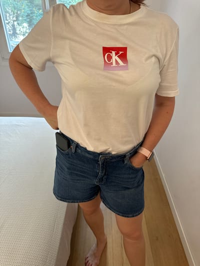 A person stands in a room, wearing a white t-shirt with a red and pink gradient logo on the chest, and denim shorts. A black smartphone is tucked into the front pocket of the shorts, and a pink smartwatch is on the person's left wrist. The person's left hand is in their pocket, and their right hand is by their side. The background shows a window with trees visible outside, a white wall, a light-colored bed, and a wooden floor. The image does not contain any information about the city of Murcia, Spain.