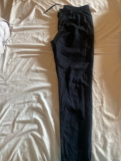 A pair of black sweatpants or joggers is laid out flat on a light-colored, possibly white or off-white, rumpled bedsheet. The pants are positioned horizontally across the frame, with the waistband and drawstring visible on the left side. The fabric appears soft and somewhat creased, suggesting a comfortable material like cotton.

On the upper left thigh area of the pants, there is a subtle, dark grey or black printed or embroidered logo and text. Although difficult to read due to low contrast, the text clearly includes the word "COURAGE" at the top. Below "COURAGE", the words "CREATIVE" and "HUMANS" can be discerned, potentially forming "COURAGE CREATIVE HUMANS". A small, circular or stylized logo appears above the word "COURAGE".

The scene is indoors, likely a bedroom, indicated by the bedsheet. The lighting is soft and even, suggesting indirect natural light, possibly from a window, casting gentle shadows and highlights on the fabric and sheet. There are no people visible, and no activity is taking place. In the bottom left corner, a small portion of another light-colored garment is partially visible.