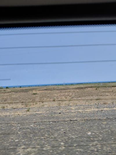 The image depicts a wide, horizontal view of a landscape, likely taken from a moving vehicle. The foreground is dominated by a rough, grey surface with visible pebbles and debris, suggesting a road or unpaved ground. In the mid-ground, dry, light brown grass and sparse bushes stretch across a flat expanse. A faint line of trees and scattered smaller bushes can be seen on the horizon, marking the transition to the background. The background is a vast, clear blue sky, with a slightly lighter hue near the horizon where it meets a distant, thin blue line that could represent water or a further expanse of land. Several thin, dark horizontal lines, possibly power lines, are visible across the sky, indicating human infrastructure. The overall impression is of an arid or semi-arid environment under a bright sky. The location is identified as Ынталы, Kazakhstan, a region known for its steppe landscapes. There are no people or animals clearly visible in the image.
