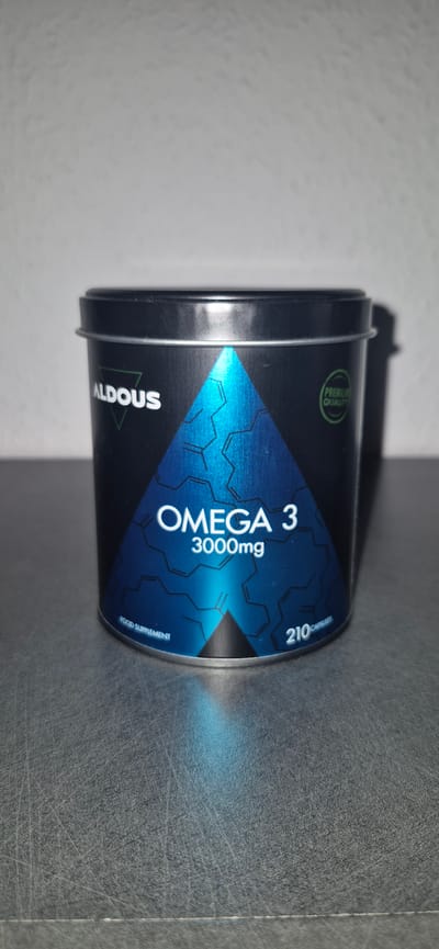 A black tin can labeled "ALDOUS OMEGA 3 3000mg" sits on a textured gray surface. The can has a blue triangular design with a molecular structure graphic. A green circular emblem on the can reads "PREMIUM QUALITY". Text at the bottom of the can says "FOOD SUPPLEMENT 210 CAPSULES". The background is a plain, textured white wall. There are no people present. The lighting casts a shadow of the can to the right. The image is taken from a slightly elevated angle, looking down at the can. The scene is indoors, likely a shelf or table. There is no indication of the city of Baden-Baden, Germany, being present in this image.