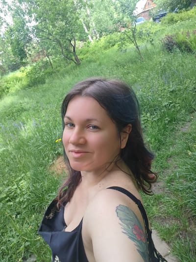 A woman is posing for a selfie in an outdoor setting. The woman has dark hair with hints of blue and red, and a tattoo on her left shoulder. She is wearing a dark top. The background consists of a grassy area with trees and foliage. In the upper part of the frame, glimpses of a house and a car can be seen. The location is Свистуха (кузяевский С/о), Russia.