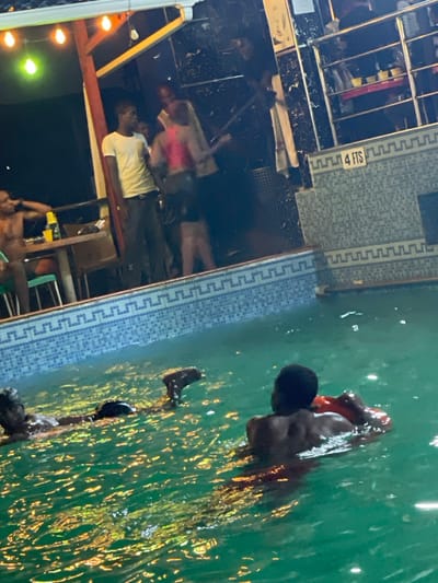 A group of people gathers near a pool, some partially submerged in the water. One person is positioned in the water, using a red floatation device. The pool's edge is marked with "4 FTS". Adjacent to the pool, people are standing or seated near tables and chairs under string lights. The scene appears to be illuminated at night.