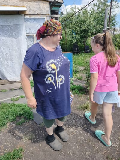 In a rural outdoor setting in Блохино, Russia, a woman and a young girl stand on a dirt path. The woman, wearing a blue t-shirt with a dandelion print and a colorful headscarf, faces the girl and appears to be speaking to her. She also wears dark shorts and grey clogs. The girl, with her hair in a ponytail, wears a pink t-shirt and light blue shorts, and is turned away from the camera, looking in the same direction as the woman. She wears light blue slip-on sandals. The background reveals a rustic wooden structure, overgrown greenery, and a blue barrel. The sky is visible, indicating it is daytime and likely sunny, with some clouds. The overall impression is a casual, everyday moment between two individuals in a simple, rural environment.