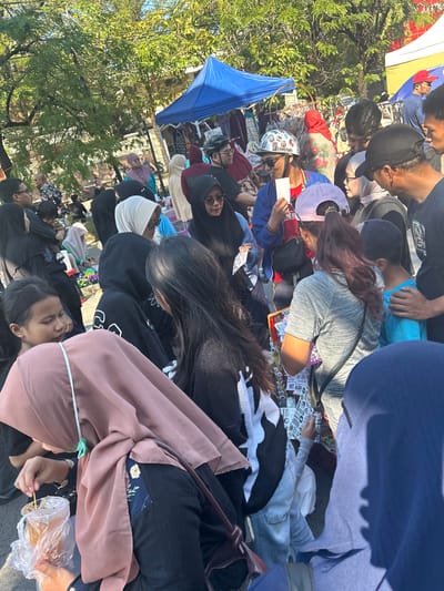 This image captures a bustling street market scene in Makassar, Indonesia, under a clear, sunny sky. The foreground is dominated by a group of people, some of whom are wearing headscarves and traditional Islamic attire, suggesting a local cultural context. A young girl in the lower left is seen holding a plastic bag with a beverage and a stick, possibly a street food item. To her right, a woman in a pink headscarf leans forward, interacting with someone off-camera. In the mid-ground, a diverse crowd browses stalls set up under blue and yellow tents. Clothing and various items are displayed for sale, indicating a commercial activity. Several individuals are looking at products, and some are engaged in conversation. The background features lush green trees and buildings, characteristic of an urban environment in Makassar. The overall atmosphere is lively and depicts a typical day at a local market.