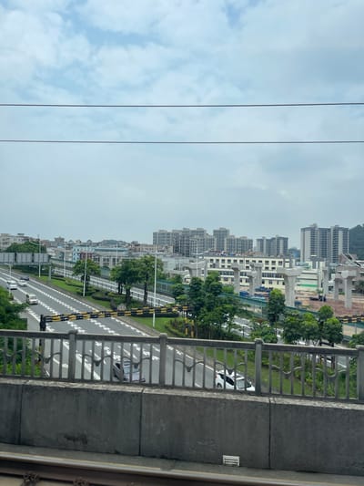 The image captures a highway in Dongguan, China, with a cityscape visible in the background. The highway is multi-laned, with cars driving on it. There are also several buildings, including residential complexes and what appears to be a construction site with large concrete pillars. Trees and green spaces line the sides of the highway, and a metal railing separates it from a pedestrian walkway or elevated structure. The sky is overcast with clouds. The overall scene depicts a developed urban area with ongoing infrastructure development.