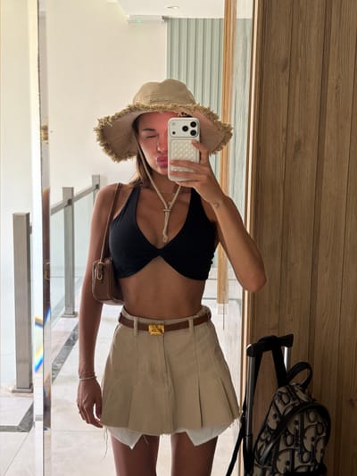 A young woman stands in a hotel hallway in Bodrum, Turkey, taking a mirror selfie. She is wearing a black bikini top, a tan bucket hat with frayed edges, and a tan pleated skirt with a white fringe hem and a brown belt with a gold buckle. She is holding a white iPhone in her right hand and has a small brown shoulder bag. A black suitcase with a patterned backpack attached to it is visible to her right. The hallway features a reflective floor and a wooden accent wall.