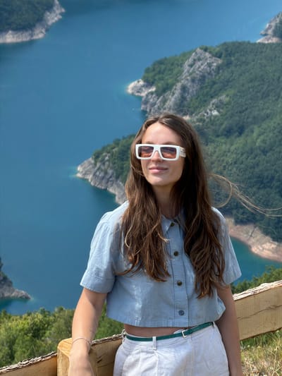 A young woman with long, brown hair and white-rimmed sunglasses stands in the foreground, looking to her left. She is wearing a light blue, cropped button-up shirt and white pants with a green belt. She has a bracelet on her left wrist. Behind her, a bright blue, winding body of water is surrounded by steep, tree-covered mountains. The scene appears to be a scenic overlook in Dubljevići, Montenegro.