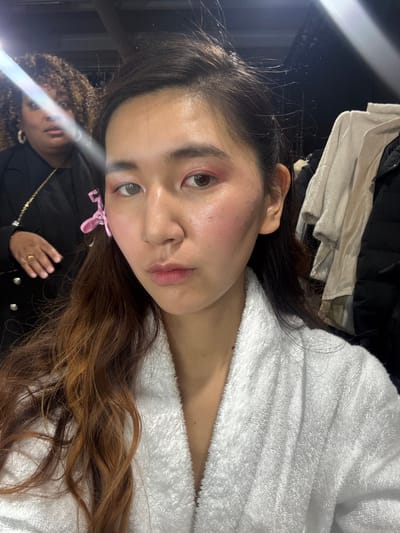 An East Asian female, appearing to be a young adult, is captured in a medium close-up selfie in Issy-les-Moulineaux, France. She wears a white terry cloth bathrobe, which is partially open at the neck. Her long, dark brown hair, with lighter reddish-brown ends, is styled down, with a pink plastic butterfly clip securing some hair on the left side of her head. Her face has visible makeup, including pink eyeshadow on both eyelids and prominent blush on her left cheekbone. Her right eye appears lighter in color than her left. She maintains a neutral expression while looking directly into the camera.

In the blurred background to the left, a second female of dark complexion with dark, curly hair is partially visible from the chest up. She wears a black blazer with gold buttons, a gold chain necklace, and a gold ring on her right ring finger. Her head is turned towards the left of the frame, and her right hand is slightly raised. A strong, diagonal light flare passes across the upper left portion of the image, partially obscuring the second female's face. To the right of the primary subject, blurred garments, including a light beige textured item and a dark puffer jacket, hang against a dark background, suggesting an indoor, possibly backstage or dressing room, environment.