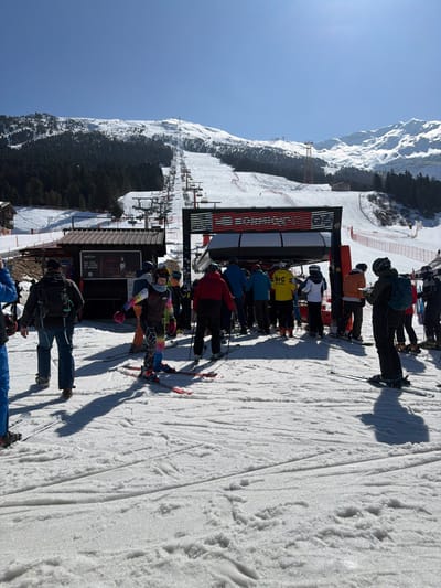 The image depicts a winter ski resort scene at the base of a chairlift in Valdisotto, Italy. Approximately 15-20 individuals, predominantly adults, are present on a snow-covered surface under clear blue skies. Most individuals are equipped with skis and poles, dressed in varied ski apparel including helmets, goggles, and colorful jackets and pants. A large cluster of skiers is positioned directly at the entry point of a modern black and red chairlift loading station, preparing to board. Several other individuals are queued behind them. Distinct shadows of the people and the chairlift structure are cast upon the foreground snow, indicating bright sunlight. To the left of the chairlift, a small wooden building with a dark roof displays a sign with "APERITIVO" and "DAL 1971." In the background, a snow-covered mountain slope ascends, featuring the chairlift line extending upwards and groomed ski trails. Evergreen forests cover portions of the lower mountain, while more rugged, snow-capped peaks are visible in the far distance. Red safety netting is present along the perimeter of the ski slope.