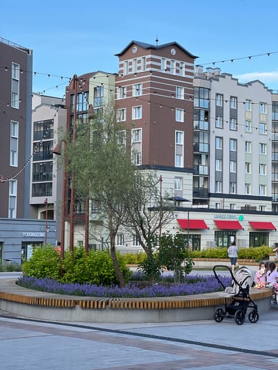 The image depicts a public square in the city of Pervomaysky, Russia, featuring modern residential buildings and a landscaped area. In the foreground, a curved wooden bench surrounds a planter filled with lush green bushes and blooming purple flowers. A baby stroller is parked to the right of the bench. In the mid-ground, a woman walks away from the camera, and a group of children are seated on the bench. Behind the planter, a tree with sparse foliage stands. The background is dominated by multi-story apartment buildings with a mix of brick and light-colored facades, characterized by numerous windows and balconies. String lights are strung across the square, adding a decorative element. Several storefronts are visible on the ground floor of the buildings, including a pharmacy with red awnings and a store labeled "30OMATAƵИH". The sky is clear and blue, indicating daytime.