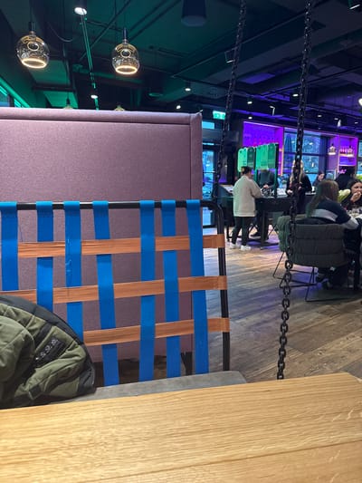 The image shows the interior of a restaurant or bar, possibly in Amstetten, Austria. In the foreground is a chair with a woven seat of blue and brown straps, and a corner of a wooden table. A green jacket rests on the chair. A partition covered in mauve fabric is visible behind the chair. Above and to the left are several spherical pendant lights. In the background, people are seated at tables. There is a display of sauce bottles. The lighting is mixed, with green and blue accents, and the floor is wood. Chains hang from the ceiling.