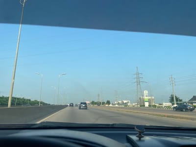 This media file captures a view from inside a vehicle traveling on a highway in Karmajiji, Nigeria. The scene is set during the daytime, under a clear blue sky, suggesting fair weather. The highway is paved and appears to be multi-lane, with a concrete barrier separating the direction of travel from oncoming traffic or another section of the road.
Several vehicles are visible on the highway, including cars and SUVs, moving in the same direction as the camera. Tall lampposts line the side of the road, indicating it is a well-lit urban or suburban area. In the background, there are numerous electricity pylons and transmission lines stretching across the horizon, a common sight in areas with significant power infrastructure.
Beyond the highway and power lines, there are some buildings, including what appears to be a multi-story structure with a sign that reads "AFDIN". Greenery, likely trees and bushes, is also present along the roadside and in the distance. The perspective suggests the photo was taken while moving, likely by a passenger or driver, as the dashboard of the car is partially visible in the foreground. There are no discernible people in the immediate foreground or background, and the overall impression is one of everyday travel on a well-developed road network.