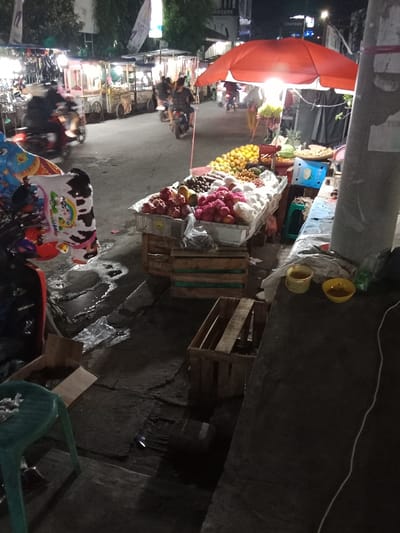The image shows a street scene in Kaliwungu, Indonesia, located at coordinates -6.961, 110.253. A fruit vendor is selling fruit from a stand on the sidewalk under a red umbrella. The fruit is arranged in wooden crates. There are two small yellow containers with a red rim in the foreground on a concrete surface. There is a green plastic chair to the left of the containers. A motorcycle is parked in the foreground with a toy on the seat. The street has a cracked pavement and a visible drain. There are several buildings to the left of the frame.