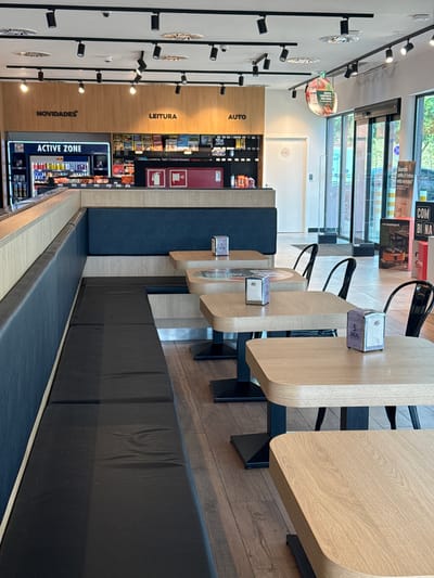 The image shows the interior of a service station shop in Oeiras, Portugal. The shop features a long, dark blue upholstered bench seat along the left side of the image, facing a row of light wood tables. Each table is set with a silver napkin dispenser. In the background, a counter area displays various products, including drinks in a refrigerator labeled "ACTIVE ZONE," magazines, and other convenience items. Above the counter, signs indicate "NOVIDADES," "LEITURA," and "AUTO." To the right, glass doors lead outside to a bright, sunlit area. A circular promotional sign hangs from the ceiling. The floor is made of dark wood planks. The overall impression is a clean and modern rest stop facility. There are no people visible in the image.