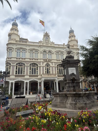 The image captures a wide-angle view of the Gabinete Literario building in Las Palmas de Gran Canaria, Spain, on an overcast day. The ornate, cream-colored building features classical architectural elements, including arched windows, decorative molding, and two distinctive domed towers. A Spanish flag flies prominently from the rooftop. In the foreground, a large, dark stone monument with a bust of a man stands beside a flower bed filled with a vibrant array of red, yellow, white, and pink flowers. To the right of the monument, a large green tree provides shade. The area in front of the building appears to be undergoing some form of setup or activity, with several ladders, scaffolding, and what look like film or event equipment present. A banner with Spanish text is visible on a lamppost to the left. Several people are casually dressed and appear to be either working or observing the scene. The overall impression is one of a public space in front of a significant cultural building, with evidence of recent or ongoing event preparation.