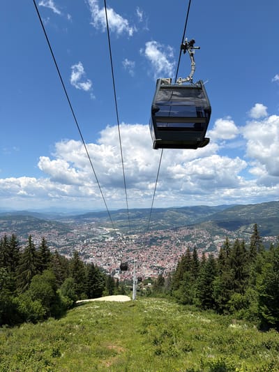 An aerial view captures the city of Sarajevo, Bosnia and Herzegovina, sprawling across a valley under a bright blue sky dotted with fluffy white clouds. In the foreground, a cable car cabin, numbered '21', hangs suspended by thick cables, descending towards the city from a grassy mountainside covered in trees. The mountainous terrain surrounding Sarajevo is visible in the distance, a hazy green against the horizon. The city itself is a dense tapestry of red-tiled roofs and urban infrastructure, with a river snaking through its center. A lower cable car station is visible on the mountainside, with another cabin en route. The scene is a panoramic depiction of urban life nestled within a natural landscape.