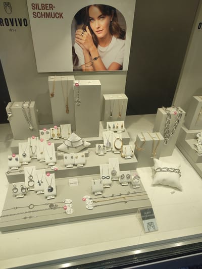 The image is a product shot of a jewelry display window, likely from a retail store in Rosenheim, Germany. The display features an assortment of silver jewelry, including necklaces, bracelets, earrings, and rings. The jewelry is presented on tiered stands and mannequins within a glass-fronted display case. A large advertisement featuring a woman modeling jewelry is visible at the top of the display, with text that includes "SILBER-SCHMUCK" and "ROVIVO 1856". Pricing tags are visible on many of the items, indicating individual prices and some discounts. The overall environment appears to be a well-lit retail setting.
