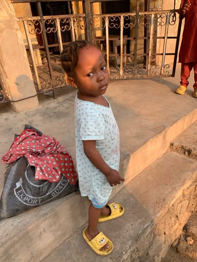 A young child wearing a light blue patterned dress lies on a concrete surface next to a patterned cloth and a black bag. The child has braided hair and wears yellow cartoon-character sandals. A metal gate sits to the side. Part of another person's legs in reddish-brown clothing and yellow sandals is also visible in the top left corner.