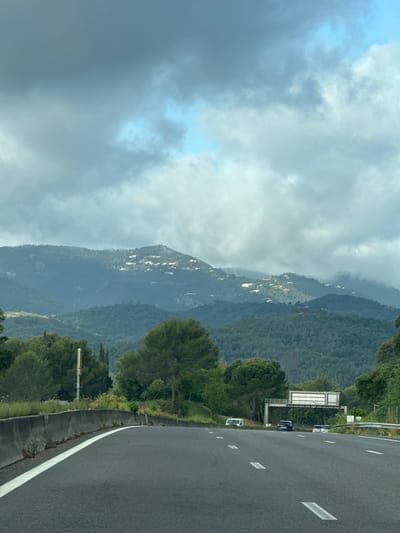 The image shows a highway winding through the mountainous terrain of Mandelieu-la-Napoule, France. The road occupies the right side of the frame and is bordered by a white line. Mountains covered in dense green vegetation rise on the left side, their peaks partially obscured by heavy, overcast clouds. Houses or buildings can be seen scattered on the slopes of the mountain. A highway sign is visible along the side of the road. A few vehicles are present on the highway.