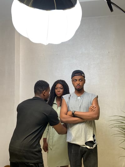 The image depicts three individuals in an indoor setting, likely a photography or video studio, illuminated by a large, white, pumpkin-shaped softbox suspended from above in the upper-left quadrant. The background is a plain, light-colored wall with a subtle texture. A green plant with long leaves is partially visible in the lower-right corner.
One male individual, with short dark hair and wearing a black short-sleeved top, is positioned with his back mostly to the viewer, facing the other two subjects. His left arm is bent, with his hand near the waist of the female subject.
A female individual, with long dark wavy hair and wearing a light green collared shirt dress, stands centrally, looking directly towards the camera with a neutral expression.
Another male individual, wearing a black baseball cap backward, a grey sleeveless t-shirt with distressed holes on the shoulders and lower left, dark trousers, a dark wristwatch on his left wrist, and a silver "AK" pendant, stands to the right with his arms crossed over his chest. He is looking towards the right side of the frame, away from the camera, with a serious expression.
There are no discernible details within the image to confirm a specific location in Moba, Nigeria.