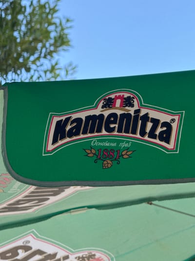 The image shows a green umbrella with the logo of "Kamenitza," a Bulgarian beer brand. The logo features the name "Kamenitza" in large, bold letters, with the text "Основана през 1881" (Founded in 1881) underneath. The umbrella is set against a backdrop of a blue sky and green foliage, suggesting an outdoor setting. In the foreground, another umbrella with a similar logo is partially visible. The scene appears to be in Belashtitsa, Bulgaria, a location known for its beer production. There are no people or specific actions depicted in the image. The focus is on the branding and the outdoor environment.
