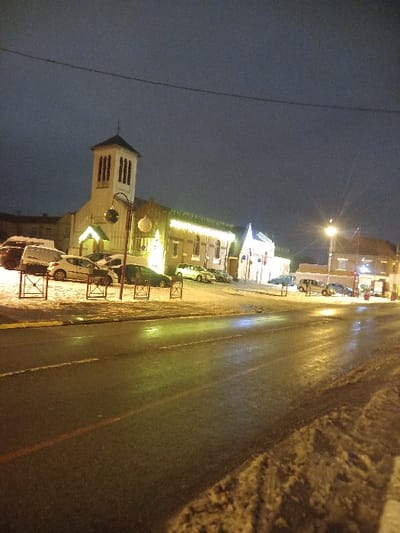 A nighttime street scene features a church with a prominent white tower to the left. The building is illuminated, with lights outlining its structure. Cars are parked along the street. A wet road reflects the glow of streetlights, creating bright streaks. The right side of the frame shows a snowy bank alongside the road. The sky is a dark, muted color.