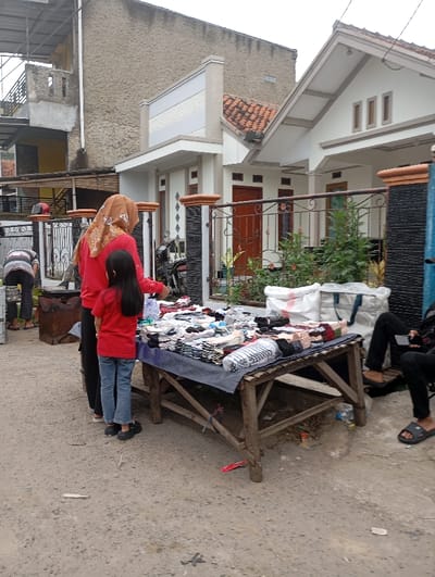 This image captures an outdoor street scene in Kutawaringin, Indonesia, under an overcast sky. In the foreground, an unpaved, gravel ground supports a makeshift vendor's stall consisting of a rustic wooden table draped with a dark grey cloth. The table displays numerous folded textile items, primarily socks or small apparel, stacked in rows, alongside a clear plastic water bottle.
Three individuals are prominently visible in the mid-ground. To the left, a person wearing a horizontal striped shirt and dark pants is partially visible from the waist down, bent over near a large, dark metal container. In the center, a woman wearing a brown headscarf (hijab), a red long-sleeved shirt, and dark pants stands with her back mostly to the viewer, examining the items on the table. Directly in front of her, a younger female, also in a red long-sleeved shirt and blue jeans, stands facing the table, observing the merchandise. To the far right, two sets of legs and feet, clad in dark pants and sandals, are partially visible, one holding a dark smartphone, suggesting individuals are seated or resting just outside the frame.
The background features residential structures. On the right, a single-story house with a white facade, a terracotta tiled roof, and a decorative metal fence is visible. A motorcycle is parked on the unpaved area beside the house's fence. To the left of this house, a multi-story building with exposed, unpainted concrete walls appears either unfinished or undergoing construction. Scattered debris, including a red wrapper and a small plastic bottle, is visible on the ground.