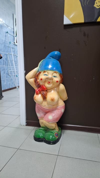A statue of a scantily clad gnome stands in what appears to be an interior space. The gnome is depicted with a blue hat, rosy cheeks, and is wearing pink polka-dotted shorts and green shoes. It is positioned next to a dark brown wall. To the left of the gnome, a tiled wall with blue square tiles is visible, along with a white drying rack. The floor is covered with light-colored square tiles. The image does not provide any specific information or context related to the city of Niukzha, Russia.