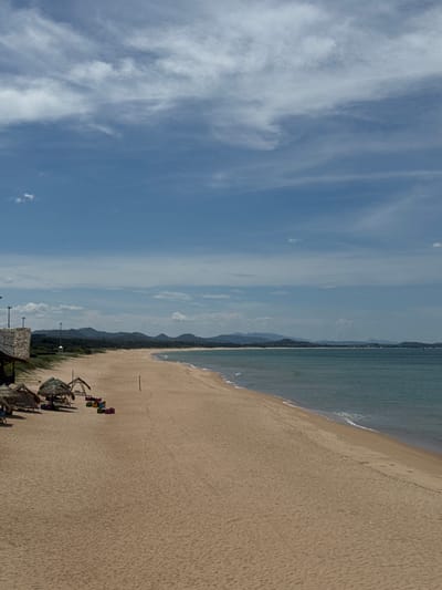 This is an aerial, sideways shot of a wide, sandy beach in Tuy Hoa, Vietnam. The beach stretches across the frame, meeting a calm, turquoise sea on the left. The sky above is a bright blue, filled with wispy white clouds. In the distance, a range of low, hazy mountains forms a backdrop along the coastline. On the sandy beach, several colorful beach chairs are arranged under thatched umbrella structures. A few people can be seen near these structures, suggesting a leisurely day at the beach. The scene is bathed in natural light, indicating it is daytime. The overall impression is one of a peaceful, tropical beach resort.