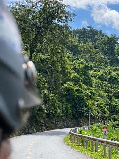 A motorcyclist's helmet, partially visible in the foreground, frames a scenic view of a winding road cutting through a lush, green mountainside. The road curves to the right, bordered by a metal guardrail and tall, vibrant green grass. A red and white marker with the numbers "H4" and "15" stands next to the road. Overhead, a clear blue sky is dotted with fluffy white clouds. The dense vegetation covering the mountains suggests a tropical or subtropical environment, consistent with the location of Thuận Hòa Commune, Vietnam, known for its verdant landscapes and mountainous terrain. The scene captures a moment of travel through a natural, picturesque setting.