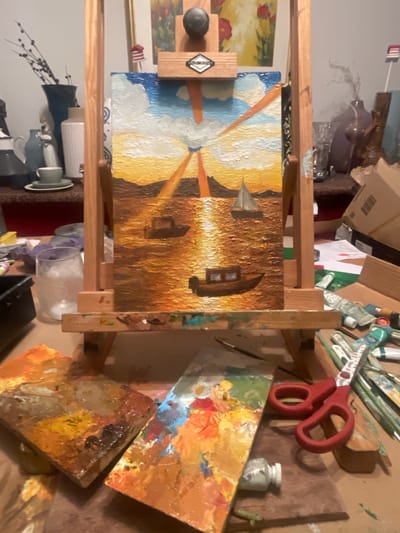 The image shows an artist's workspace. An easel holds a painting depicting a sunset with golden light reflecting on water, with a sailboat and buildings silhouetted against the bright light. Various art supplies, including paint tubes, brushes, scissors, and palettes covered with dried paint, are scattered around the easel on what appears to be a cluttered table. A small jar sits near one of the palettes. Below the easel, there are a few cups, a vase, and a jar. The location is difficult to determine based solely on the image, and the city of Maidstone, United Kingdom, cannot be inferred from the visual content.