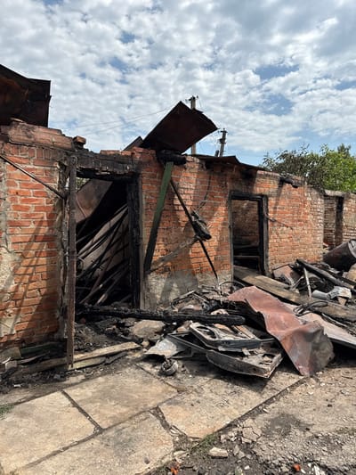 The image depicts a severely fire-damaged building located in Nedryhailiv, Ukraine. The structure, appearing to be single-story, is primarily constructed of red brick, which exhibits extensive charring and soot discoloration, particularly around openings and at the top of the walls. The roof has largely collapsed or burned away, with twisted and blackened corrugated metal sheets visibly deformed and scattered, some angled upwards against the sky.

Multiple openings, resembling doorways or windows, are present along the facade. The leftmost opening reveals a chaotic interior filled with numerous charred, collapsed wooden beams and structural elements. Another central opening shows a dark, debris-filled void.

The ground in the foreground is covered with cracked and stained concrete paving slabs directly in front of the building, transitioning to rough earth and a significant accumulation of debris. This debris includes charred wooden planks and fragments, twisted and oxidized metal sheets, and various unidentifiable distorted metal objects, some resembling parts of a vehicle or machinery, such as a possible car door frame and bicycle components. No individuals are present in the scene. The sky above is partially cloudy with white and grey cumulus clouds, and green foliage from trees is visible in the background. The overall scene is one of significant destruction and disarray.