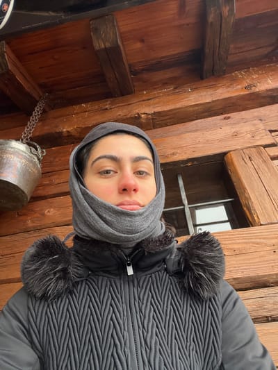 The image displays a close-up, chest-up view of a young adult female captured in a selfie in Valtournenche, Italy. She wears a grey balaclava covering her head, ears, and lower face, exposing her eyes, nose, and mouth; her nose appears reddened. She is dressed in a black puffer jacket featuring a textured chevron pattern on the torso and dark faux fur trim visible on both shoulders. The background consists of a rustic wooden log structure with prominent horizontal planks and overhead wooden beams. To the upper left, an aged, oxidized copper pot hangs from a metal chain attached to an overhead beam. To the right, a small window with a vertical metal bar is partially visible, reflecting a bright exterior, consistent with a cold, outdoor environment.